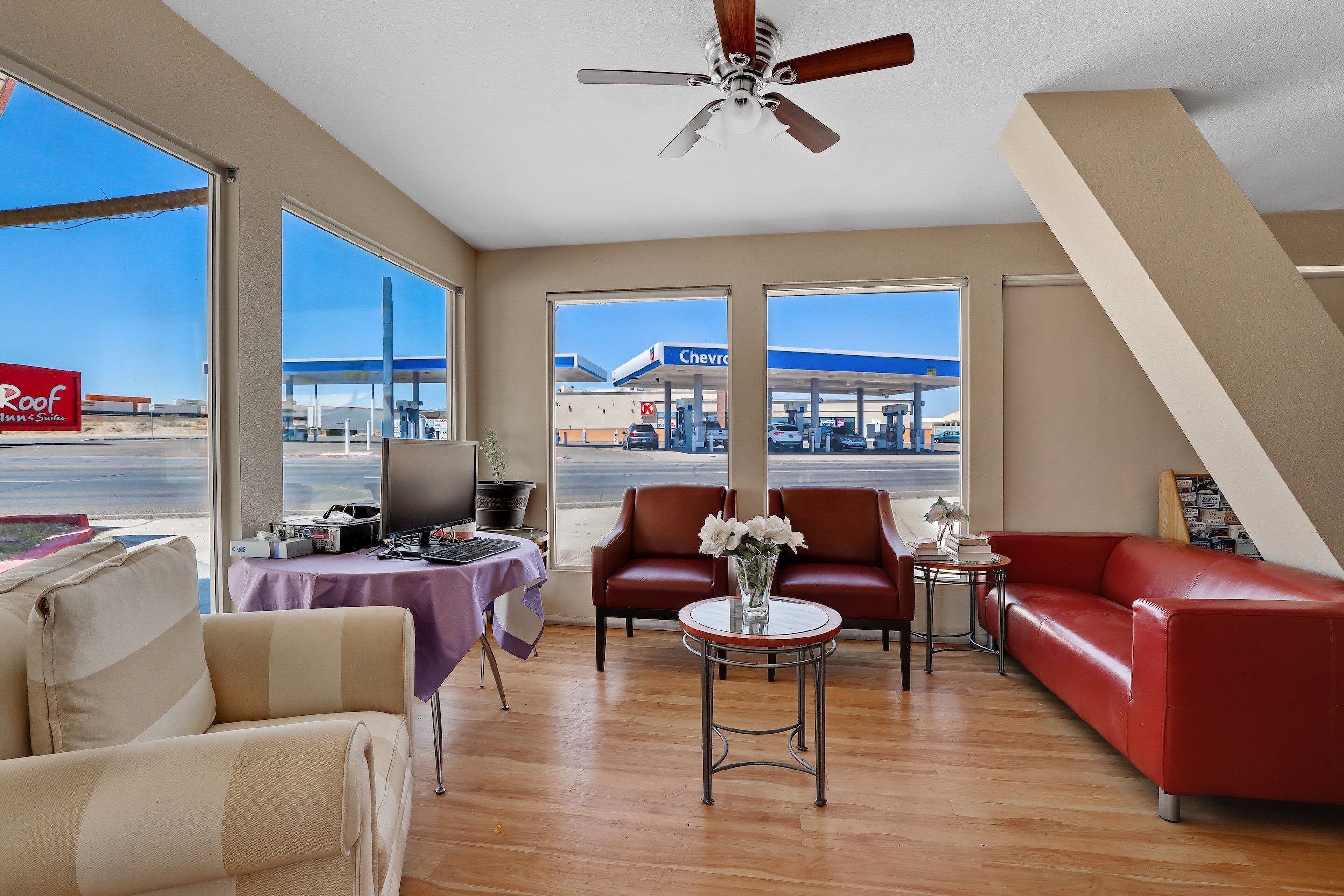 A modern living room with a red leather sofa, a beige sofa, and a round coffee table with a vase of flowers. Large windows offer a view of a Chevron gas station.
