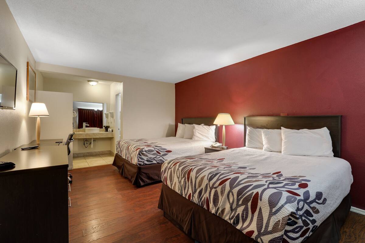 A hotel room with two beds, a desk, a mirror, and a bathroom visible through an open door.