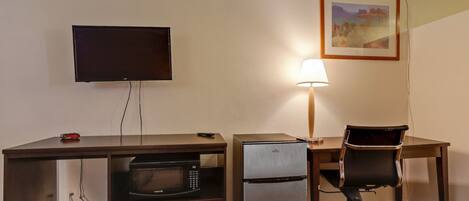 A hotel room with a desk, a television, a refrigerator, a chair, and a lamp.