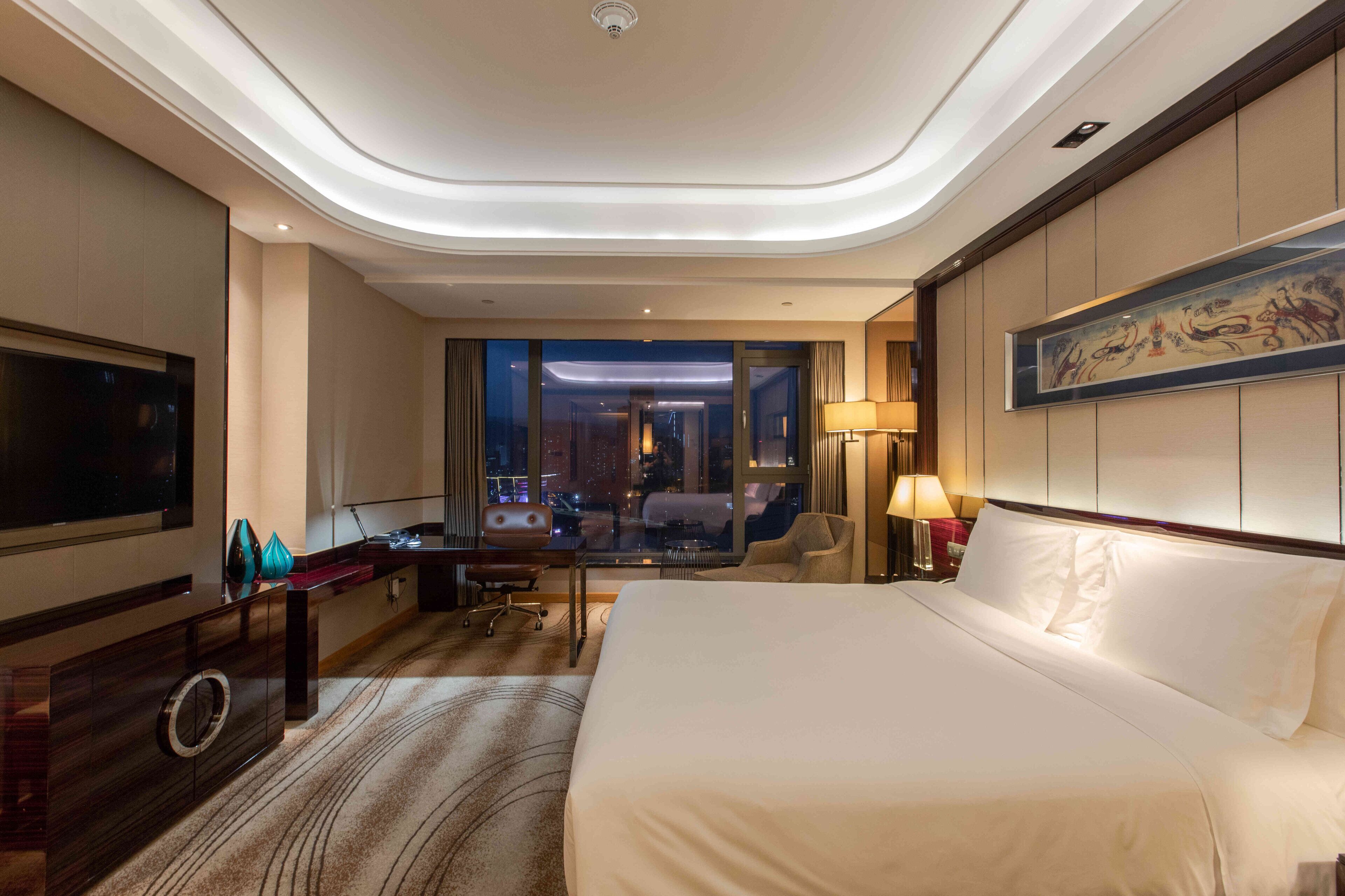 A hotel room with a large bed, a desk, a chair, a TV, and a view of the city at night.