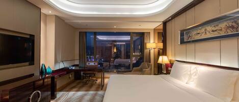 A hotel room with a large bed, a desk, a chair, a TV, and a view of the city at night.