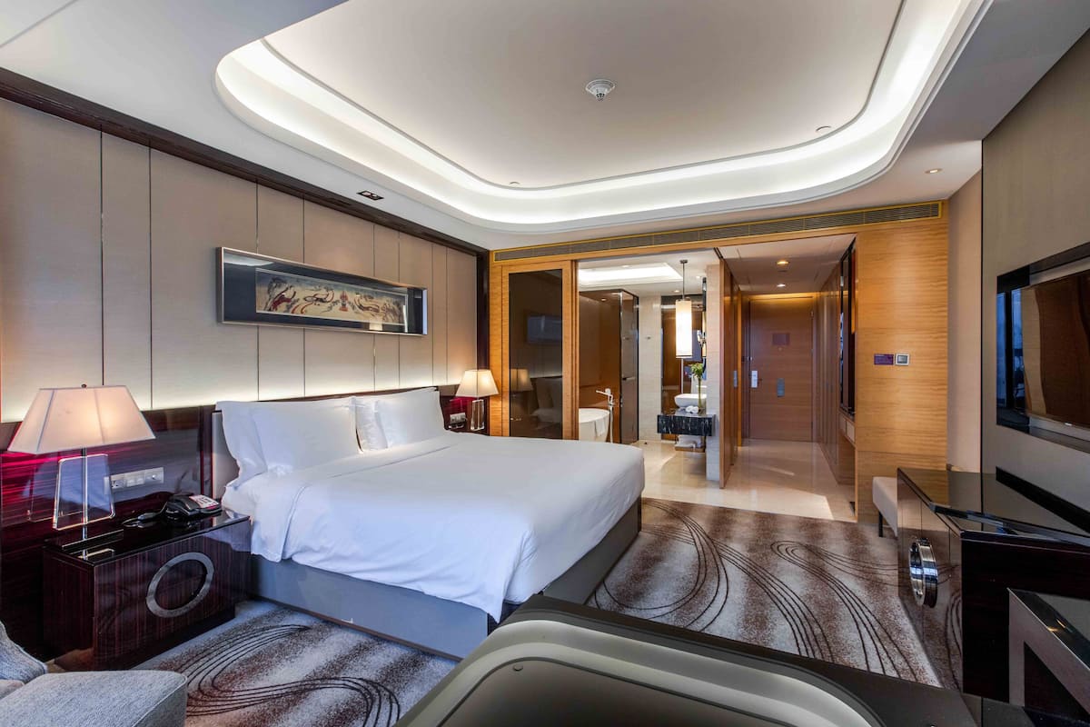 Une chambre d’hôtel moderne avec un grand lit, des lampes de chevet, un téléviseur à écran plat, un bureau avec une lampe et une salle de bain visible à travers une porte ouverte.