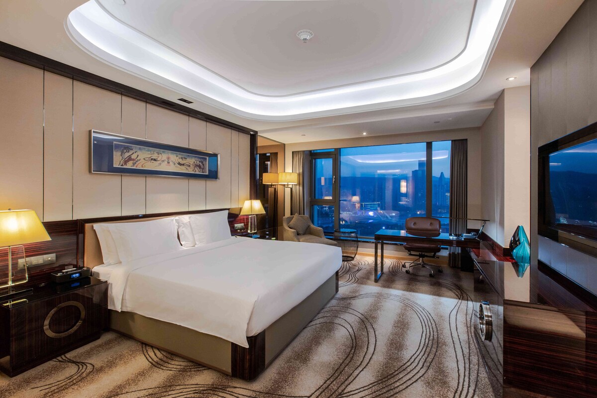 A modern hotel room with a large bed, a desk, a chair, and a view of the cityscape.
