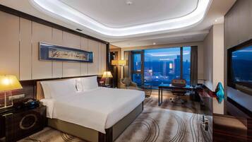 A hotel room with a large bed, a desk, a chair, a TV, and a view of the cityscape.