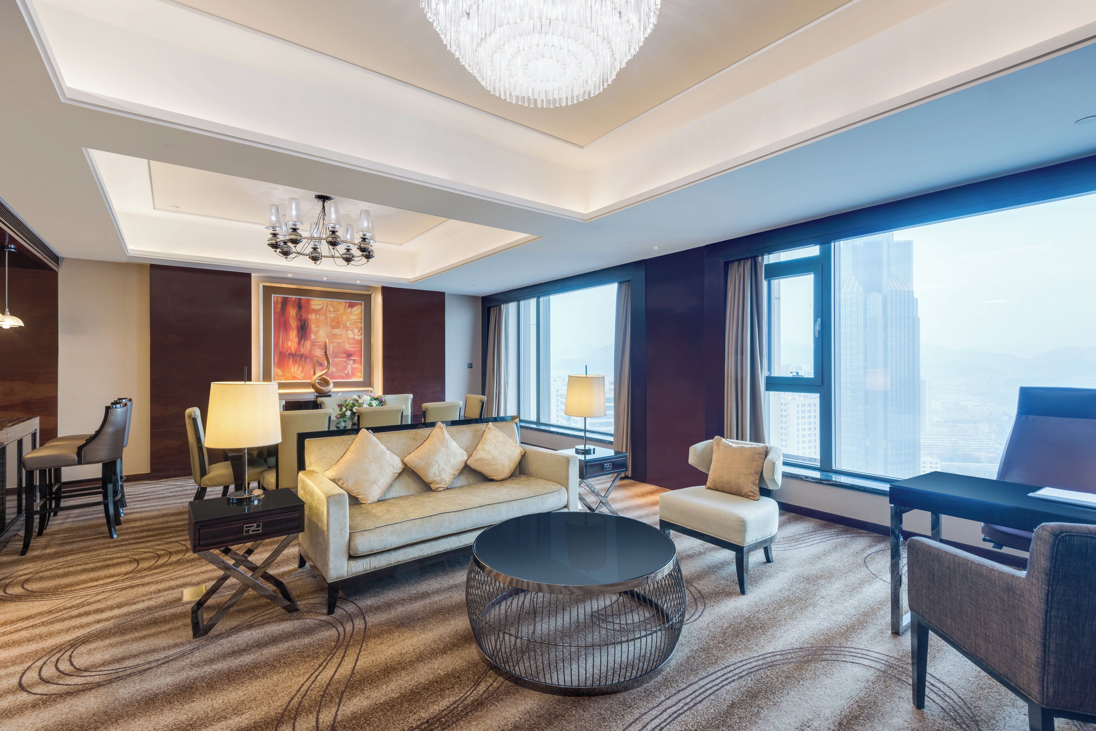 A modern hotel room with a sofa, armchairs, a coffee table, a dining area, and a large window offering a city view.