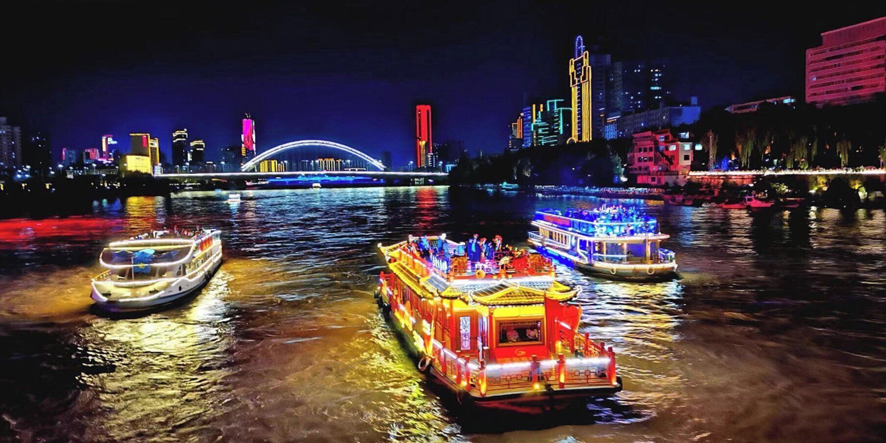 Escena nocturna de un río con barcos iluminados y un puente al fondo.