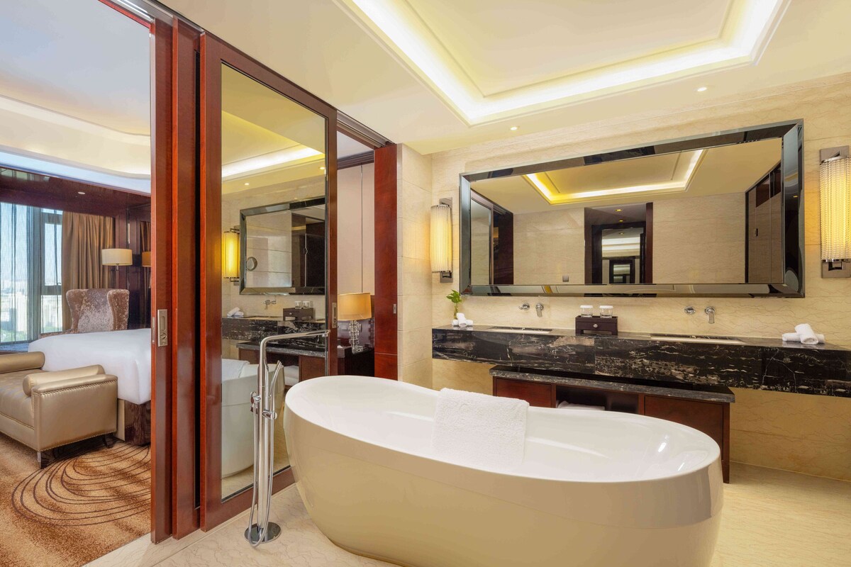 A modern hotel bathroom with a large freestanding bathtub, a double vanity with a black marble countertop, and a large mirror above the sink.