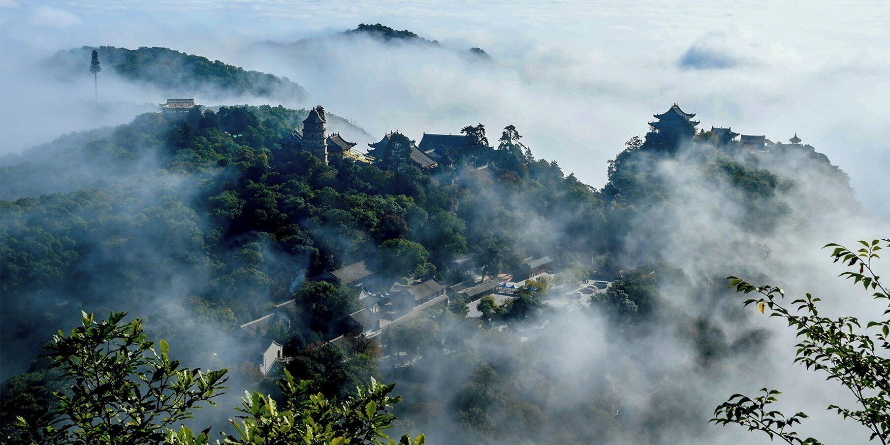 A mountainous landscape with traditional architecture, including pagodas and temples, surrounded by dense forests and enveloped in mist.