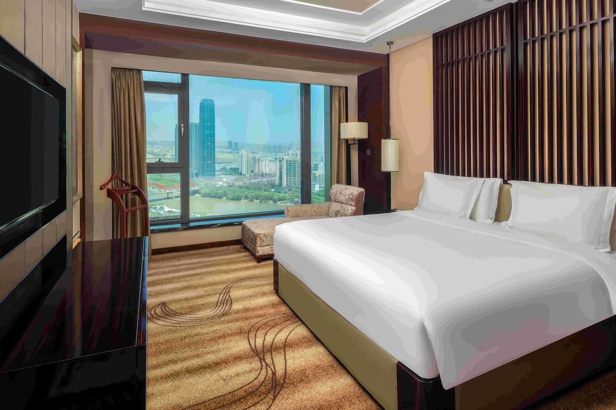 Une chambre d’hôtel avec un grand lit, une télévision, une chaise, une lampe et une vue sur le paysage urbain, visible à travers une grande fenêtre.