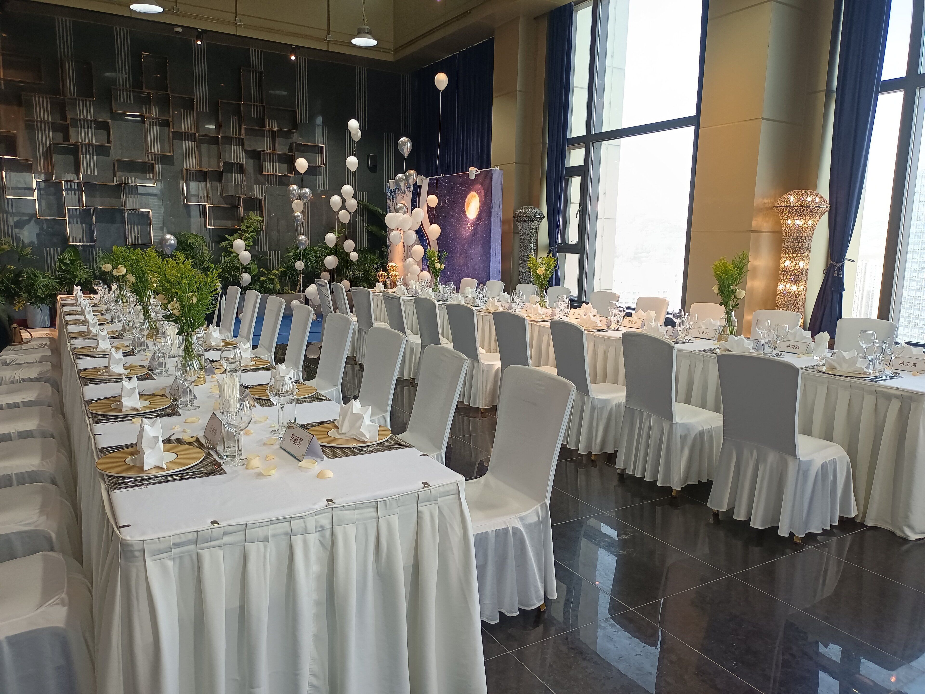 Un salón de banquetes con largas mesas dispuestas para un evento formal, con tarjetas de nombres, cristalería y centros de mesa florales.
