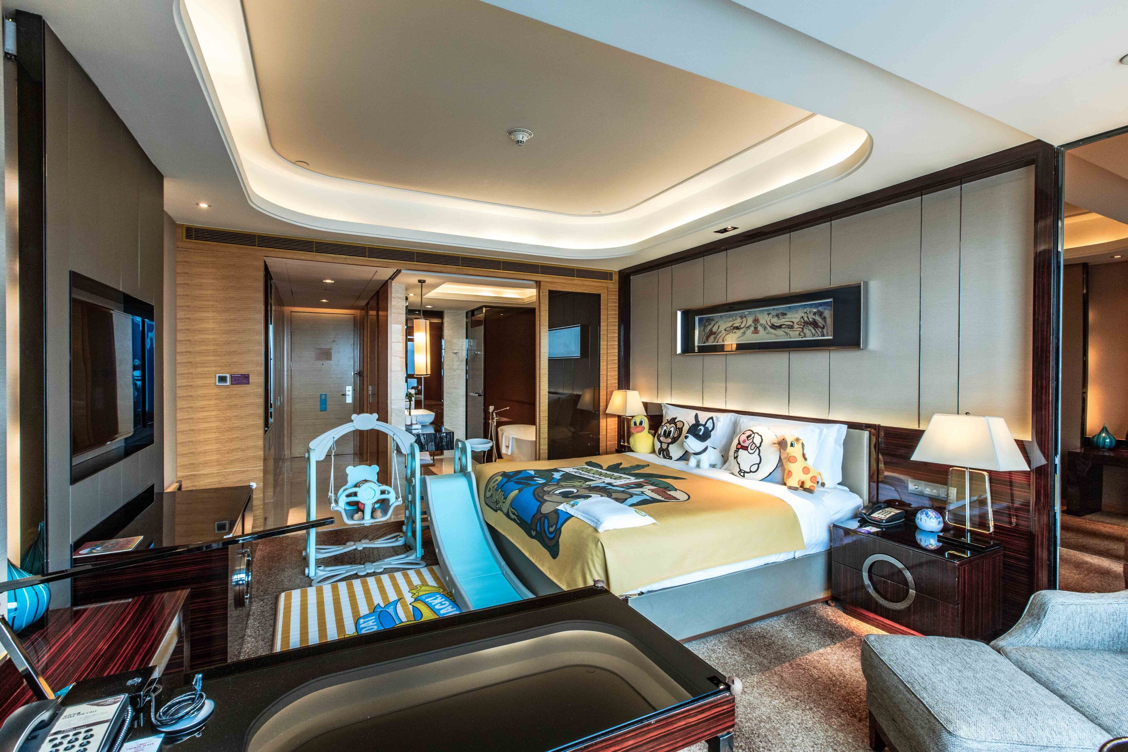 A modern hotel room with a large bed, a sitting area with a sofa, a desk with a computer, a TV, and a bathroom with a bathtub.