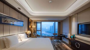 Une chambre d’hôtel moderne dotée d’un grand lit, d’un bureau avec une chaise, d’une télévision et offrant une vue sur le paysage urbain grâce à de grandes fenêtres.