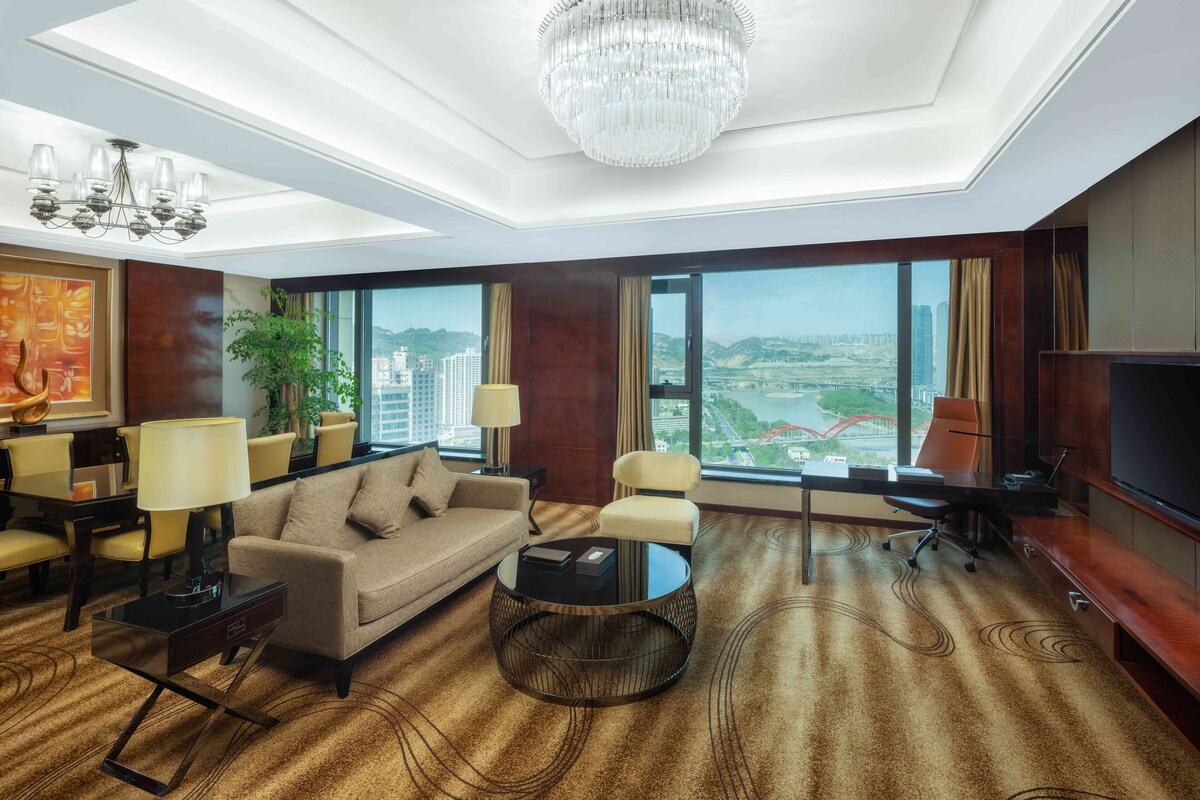 A modern hotel room with a large window offering a city view, a chandelier, a sofa, a coffee table, and a TV.