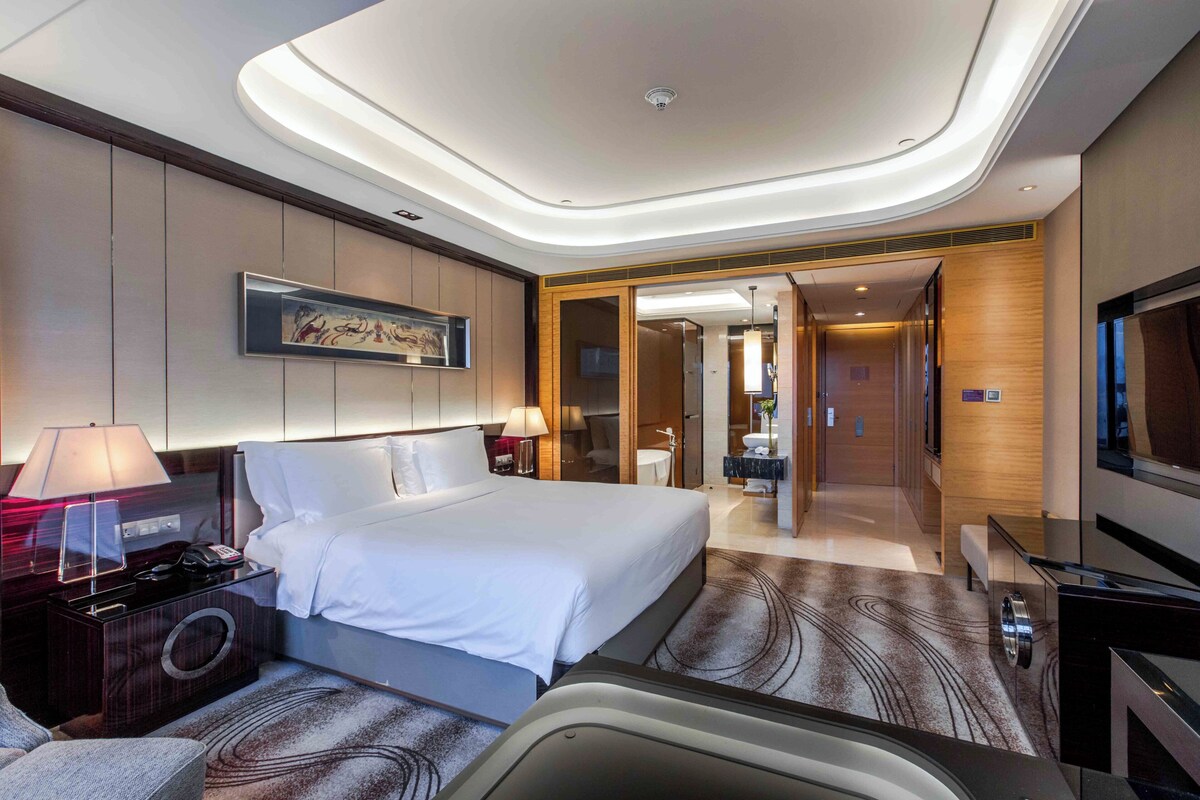 A modern hotel room with a large bed, bedside lamps, a flat-screen TV, a desk with a lamp, and a bathroom visible through an open door.