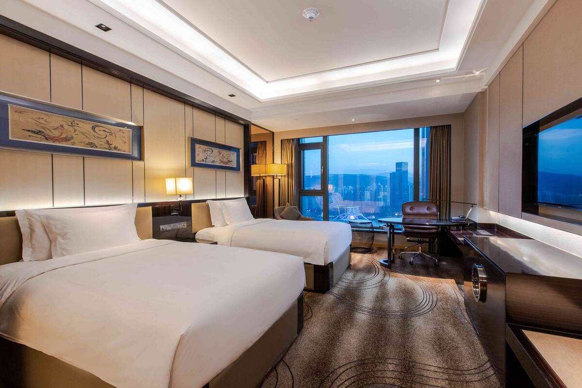 Une chambre d’hôtel avec deux lits, un bureau, une chaise, une grande fenêtre donnant sur la ville et un téléviseur à écran plat.