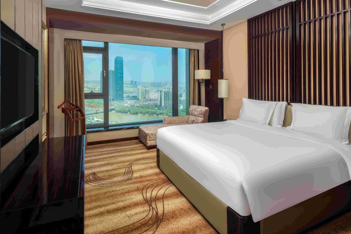 A hotel room with a large bed, a TV, a chair, a lamp, and a view of a cityscape through a large window.