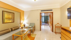 Una habitación de hotel moderna con sofá, una mesa de centro de vidrio, una silla, televisor y cómoda de madera.