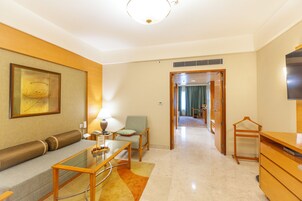 Una habitación de hotel moderna con sofá, una mesa de centro de vidrio, una silla, televisor y cómoda de madera.