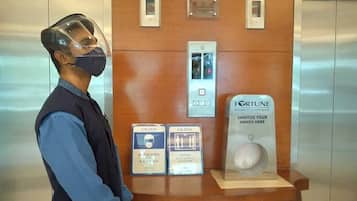 Una persona con máscara protectora y uniforme de pie frente a un mostrador de madera con dispensador de gel antibacterial y señales de precaución.