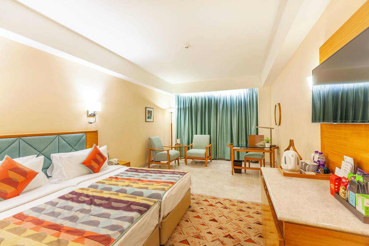 A hotel room with a bed, two chairs, a desk, a TV, and a window with curtains.