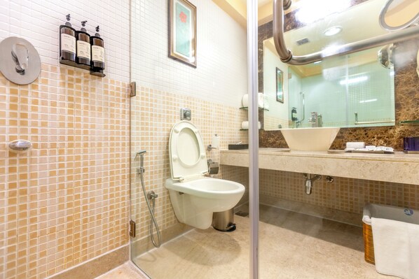 Un baño con inodoro, lavamanos y ducha. Hay botellas de jabón para manos y un dispensador de toallas en la pared.
