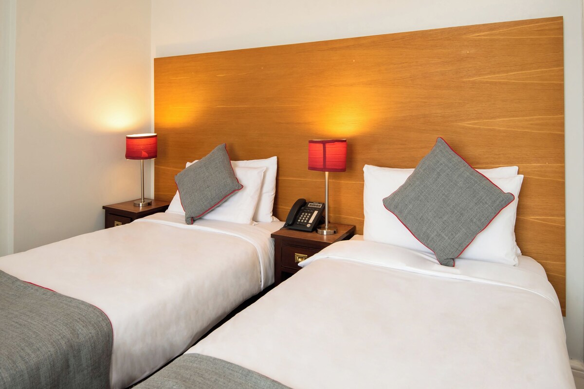 Habitación de hotel con dos camas, cada una con cabecero de madera, lámparas de noche y teléfono en la mesita.