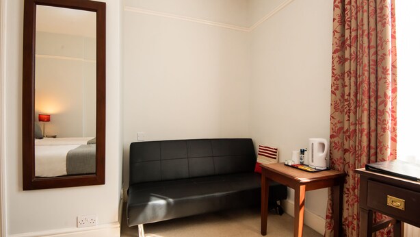 Une chambre d’hôtel avec un canapé en cuir noir, une table d’appoint en bois avec une bouilloire, un miroir et une fenêtre avec des rideaux.