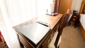 Une chambre d’hôtel avec un bureau en bois, un classeur noir portant l’inscription « The Regency Bristol Hotel – Bar Restaurant », une télécommande, un plateau avec une cafetière, un téléphone et une table de chevet avec une lampe.