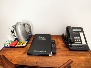 Una mesa de madera con un folleto informativo para huéspedes, un control remoto, un teléfono negro, un hervidor plateado y una bandeja con varios objetos, incluyendo una taza blanca y paquetes de comida.