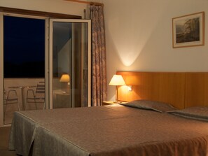 A hotel room with a bed, a nightstand with a lamp, a window with curtains, and a view of the outside. - UP Pateirahotel (Agueda)