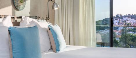 A hotel room with a bed, pillows, a lamp, and a view of a cityscape through large windows.