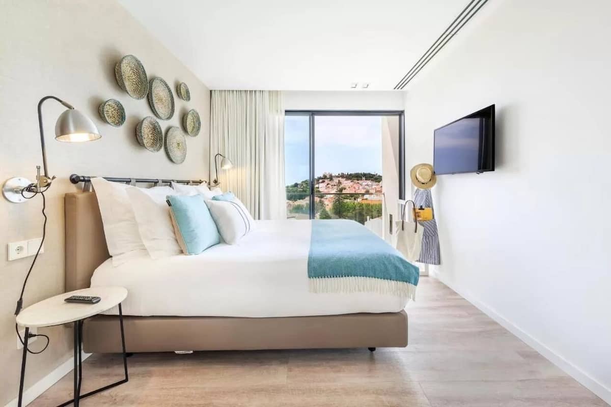 A modern bedroom with a large bed, bedside lamps, a TV, and a view of a coastal landscape.