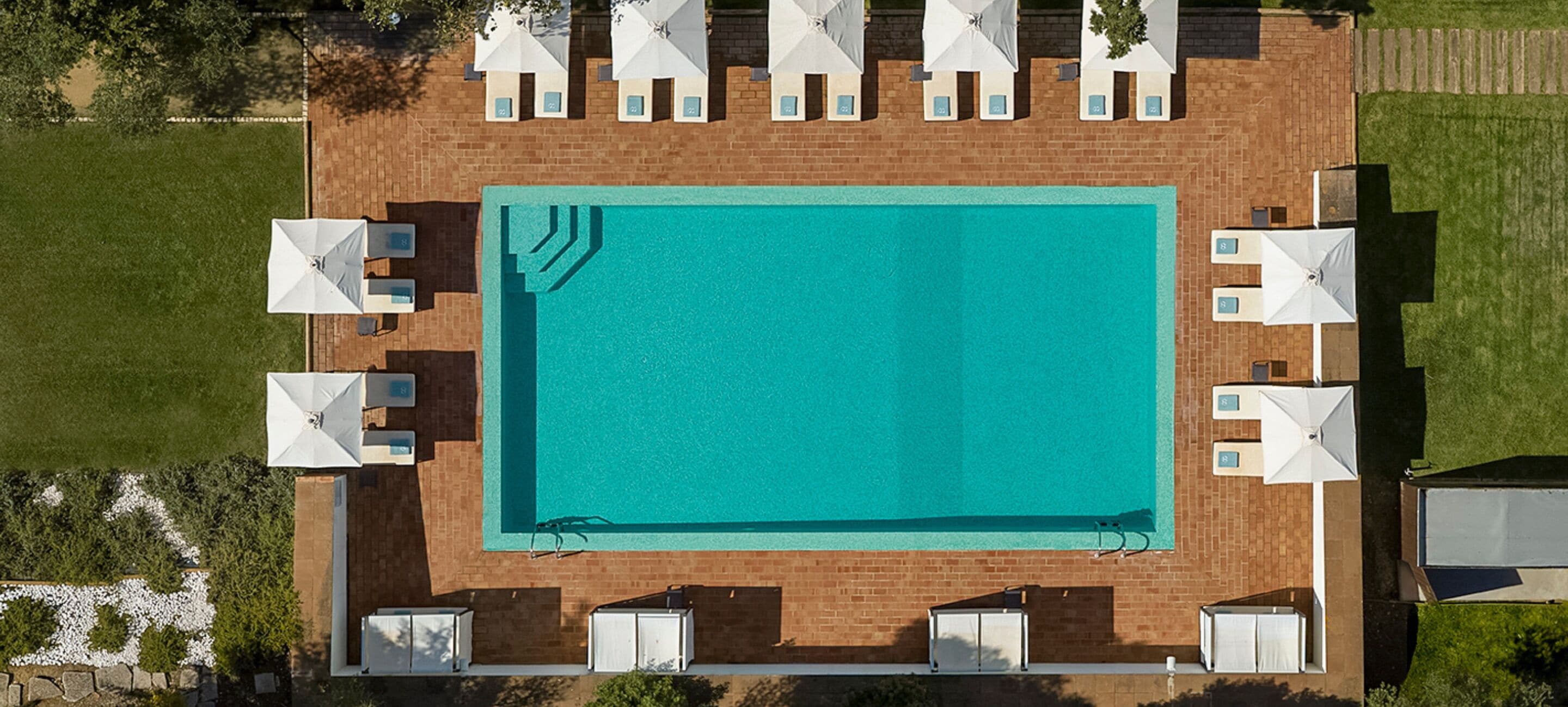 An aerial view of a rectangular swimming pool with a tiled deck, surrounded by white umbrellas and lounge chairs.