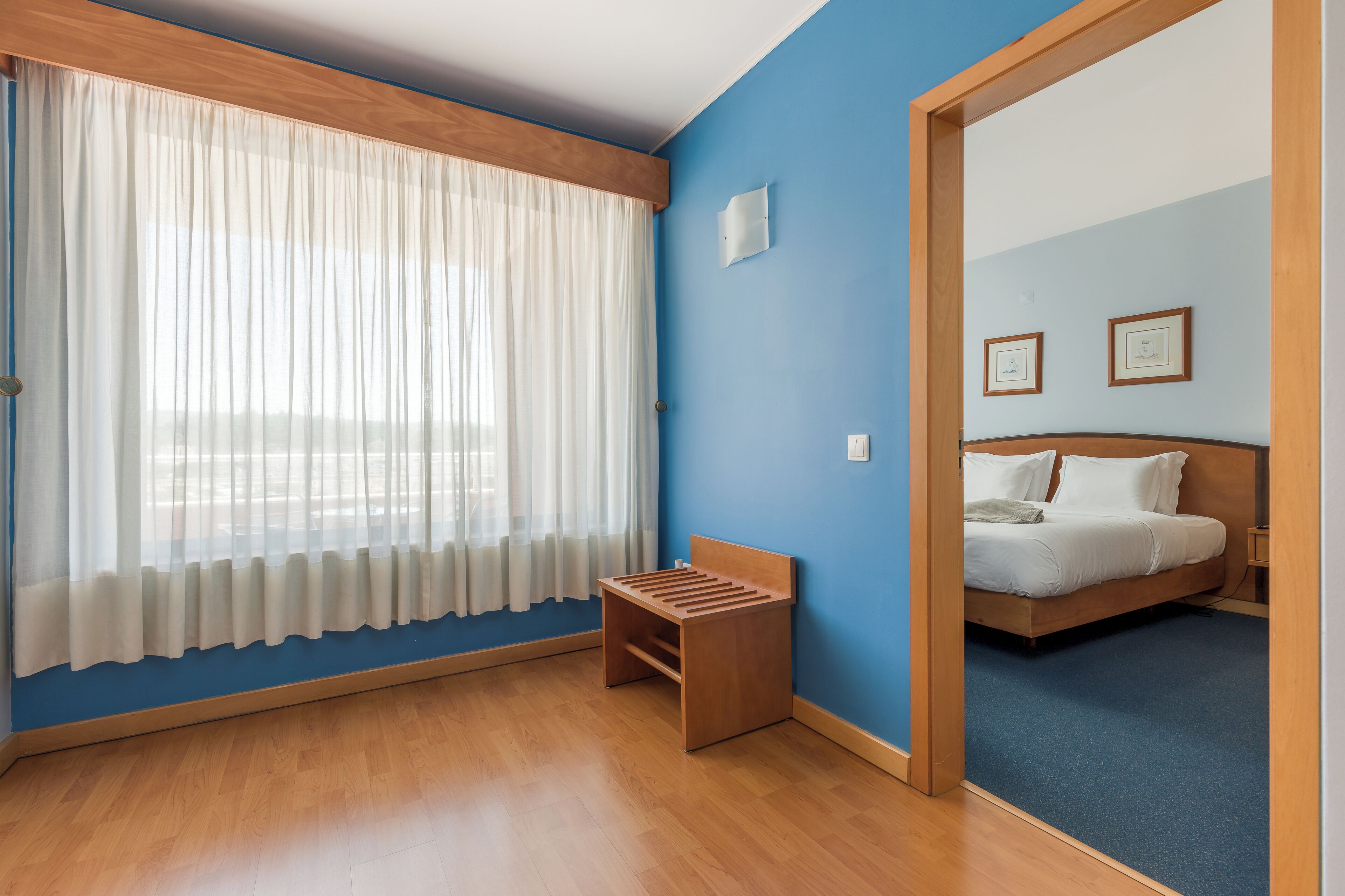 A room with a bed, a small wooden bench, a window with sheer curtains, and a blue wall.
