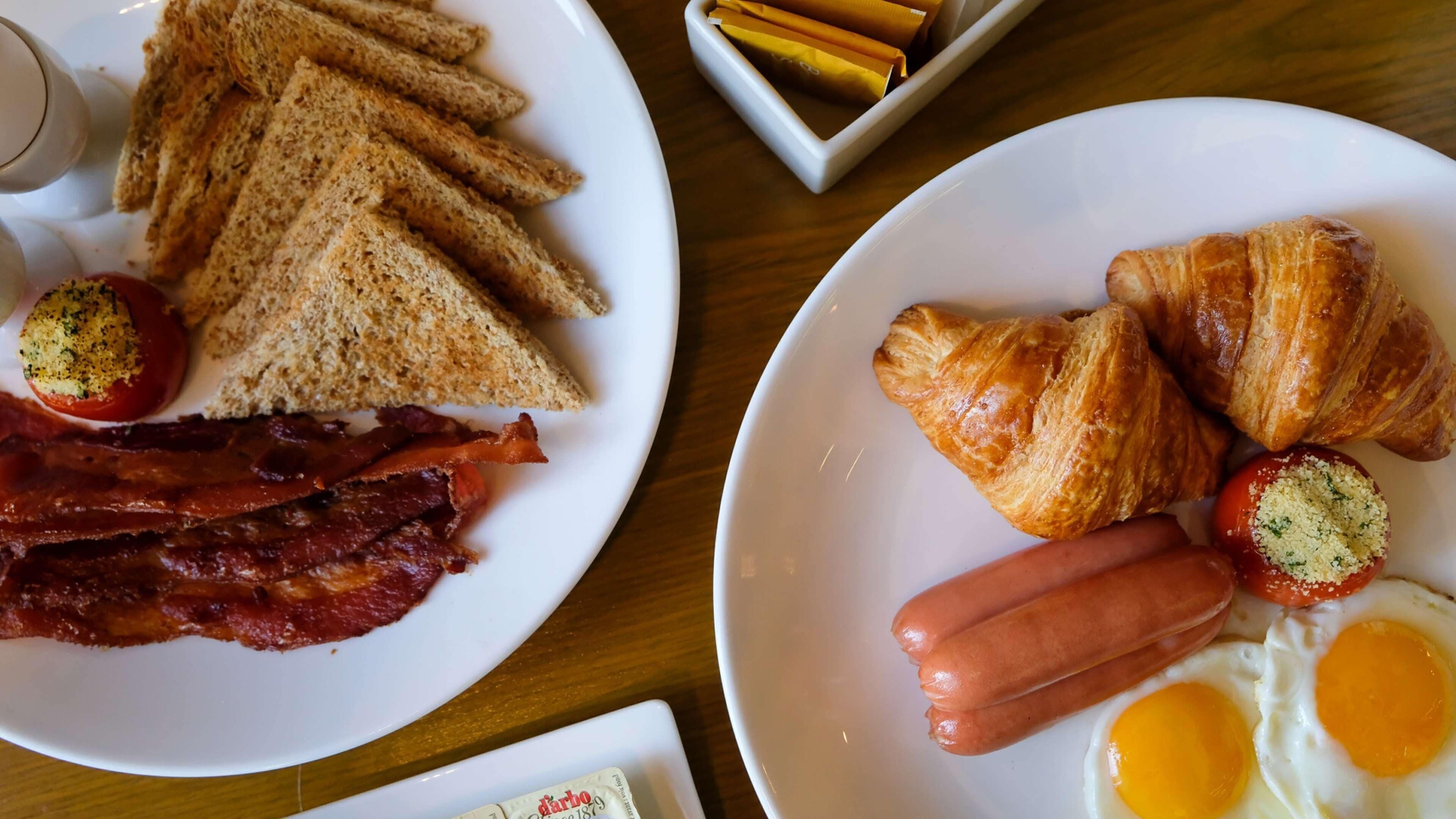 Two plates of breakfast food, including toast, bacon, sausages, eggs, and croissants.