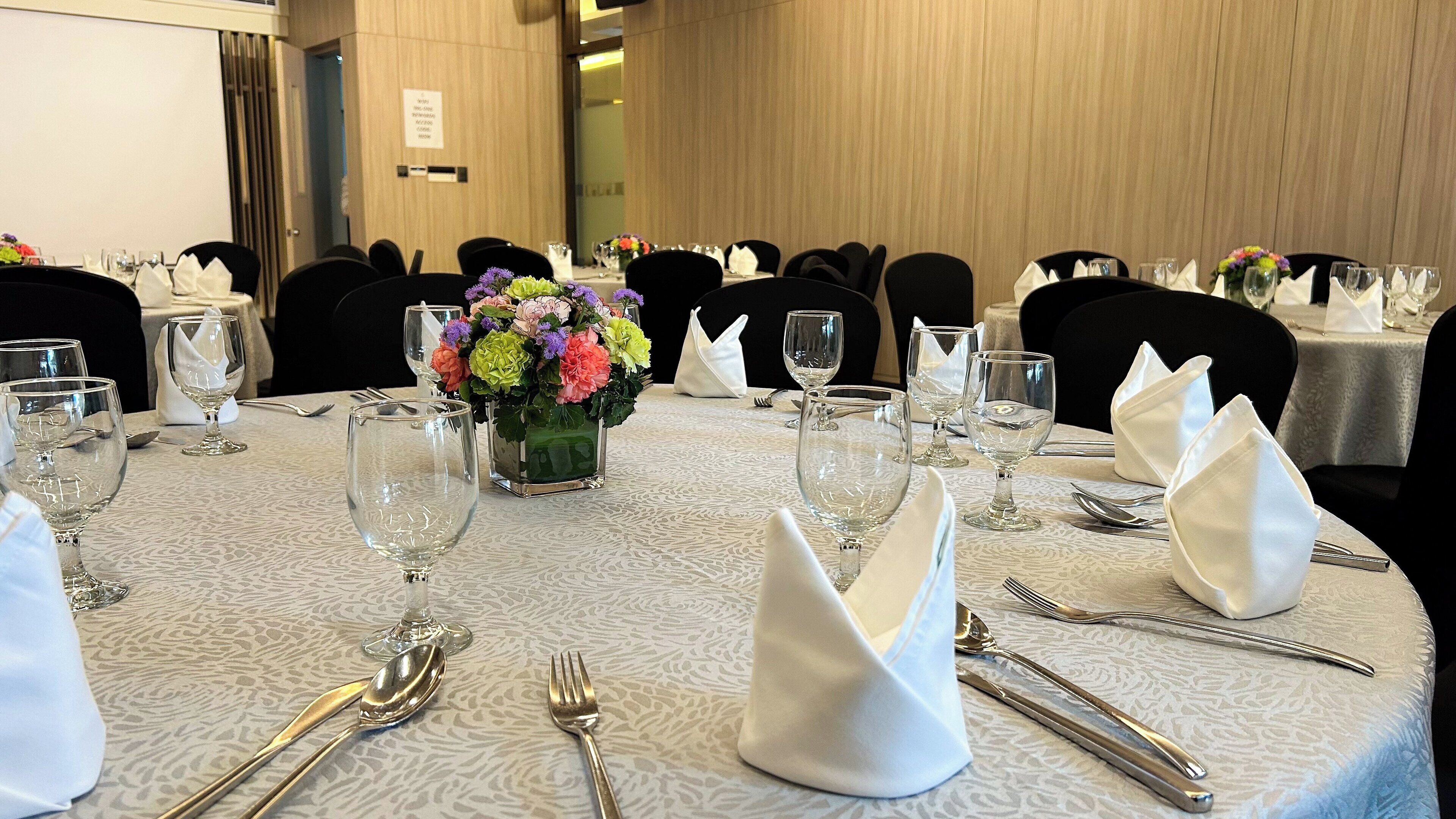 A conference room with round tables set for a formal event, complete with place settings, wine glasses, and a floral centerpiece.