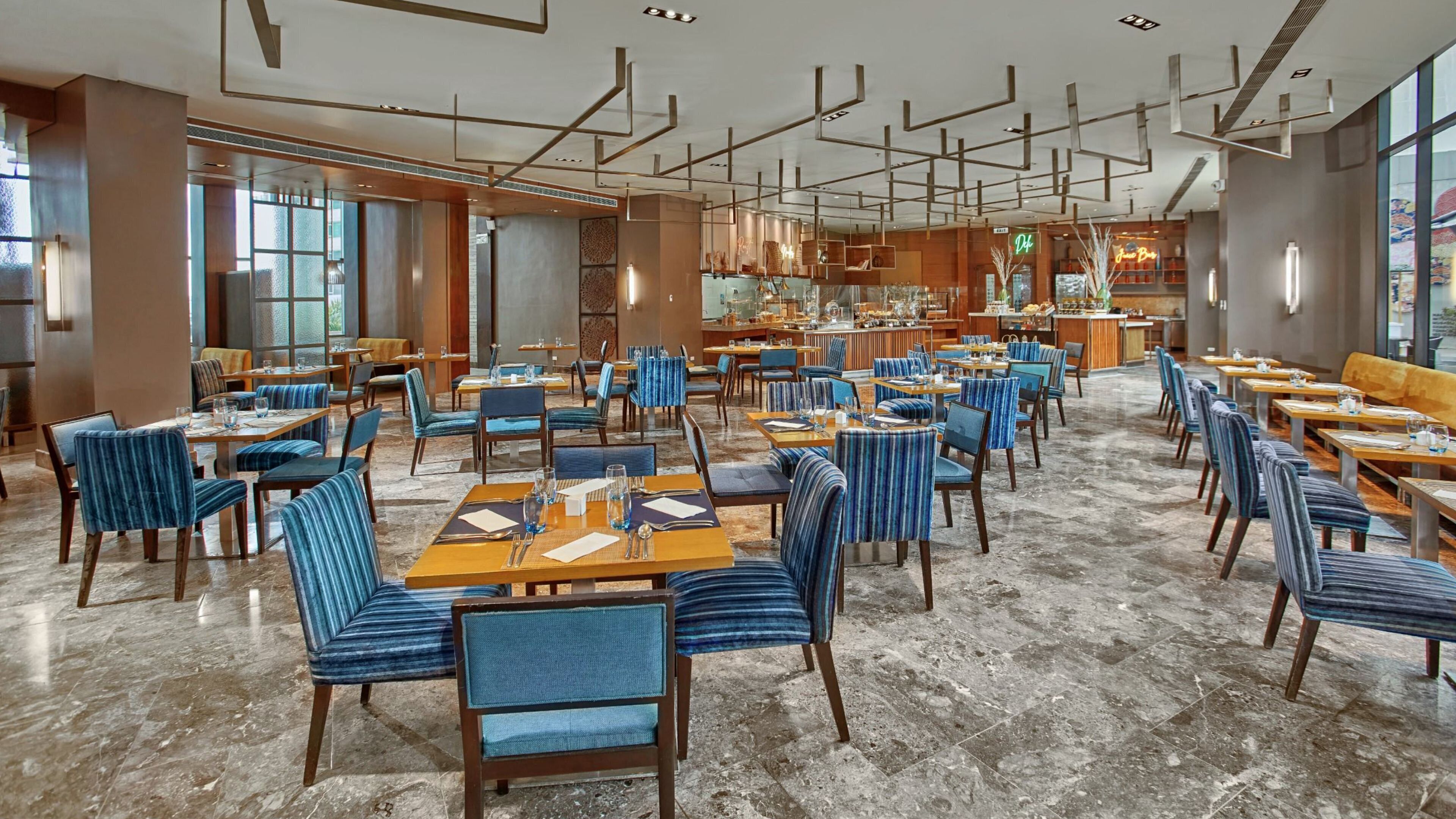 Un vaste espace de restauration dans l’hôtel, avec de nombreuses tables et chaises, un bar central et de grandes fenêtres laissant entrer la lumière naturelle.