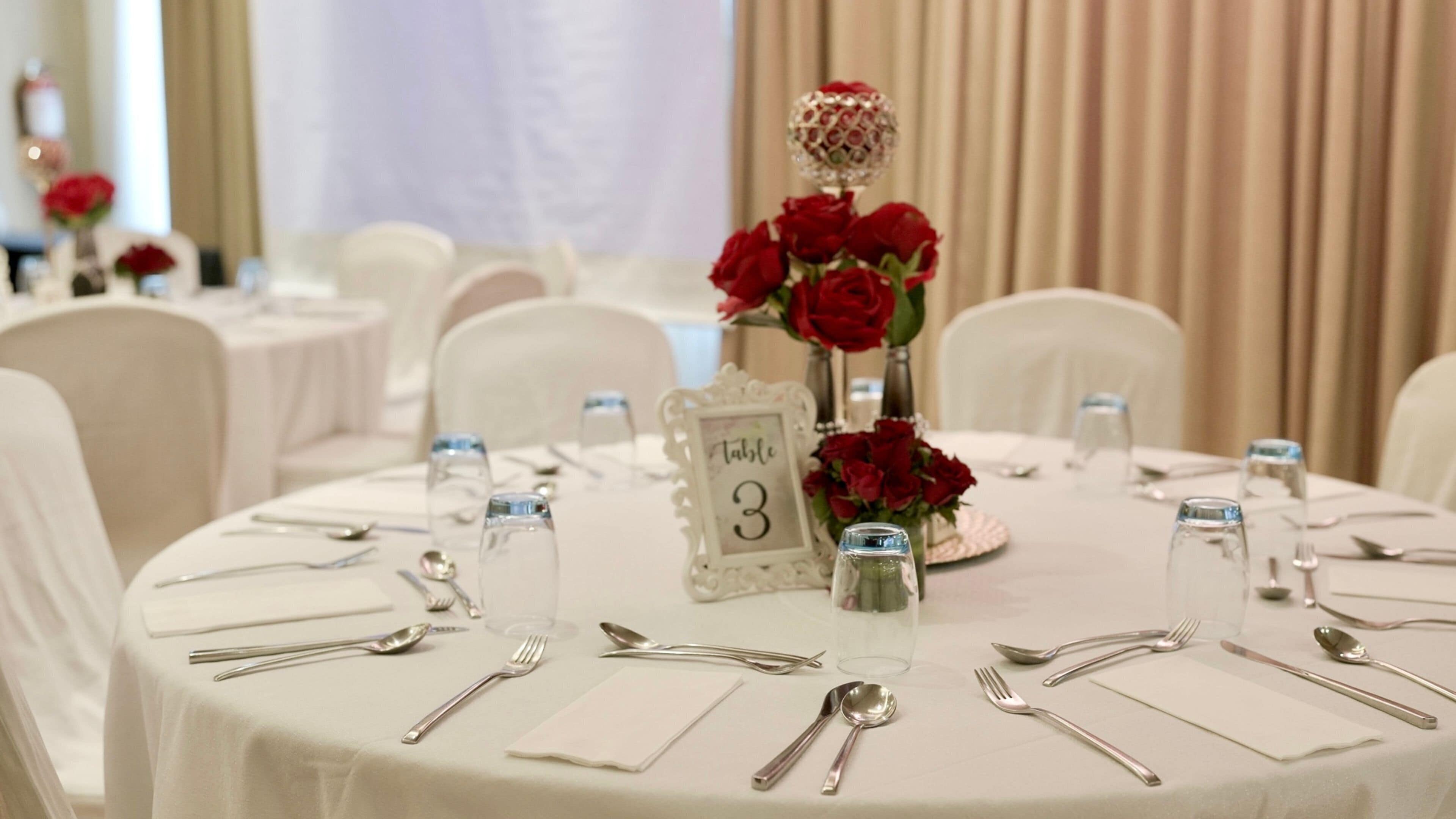 A round table with a white tablecloth, numbered table sign, and red roses in a vase.