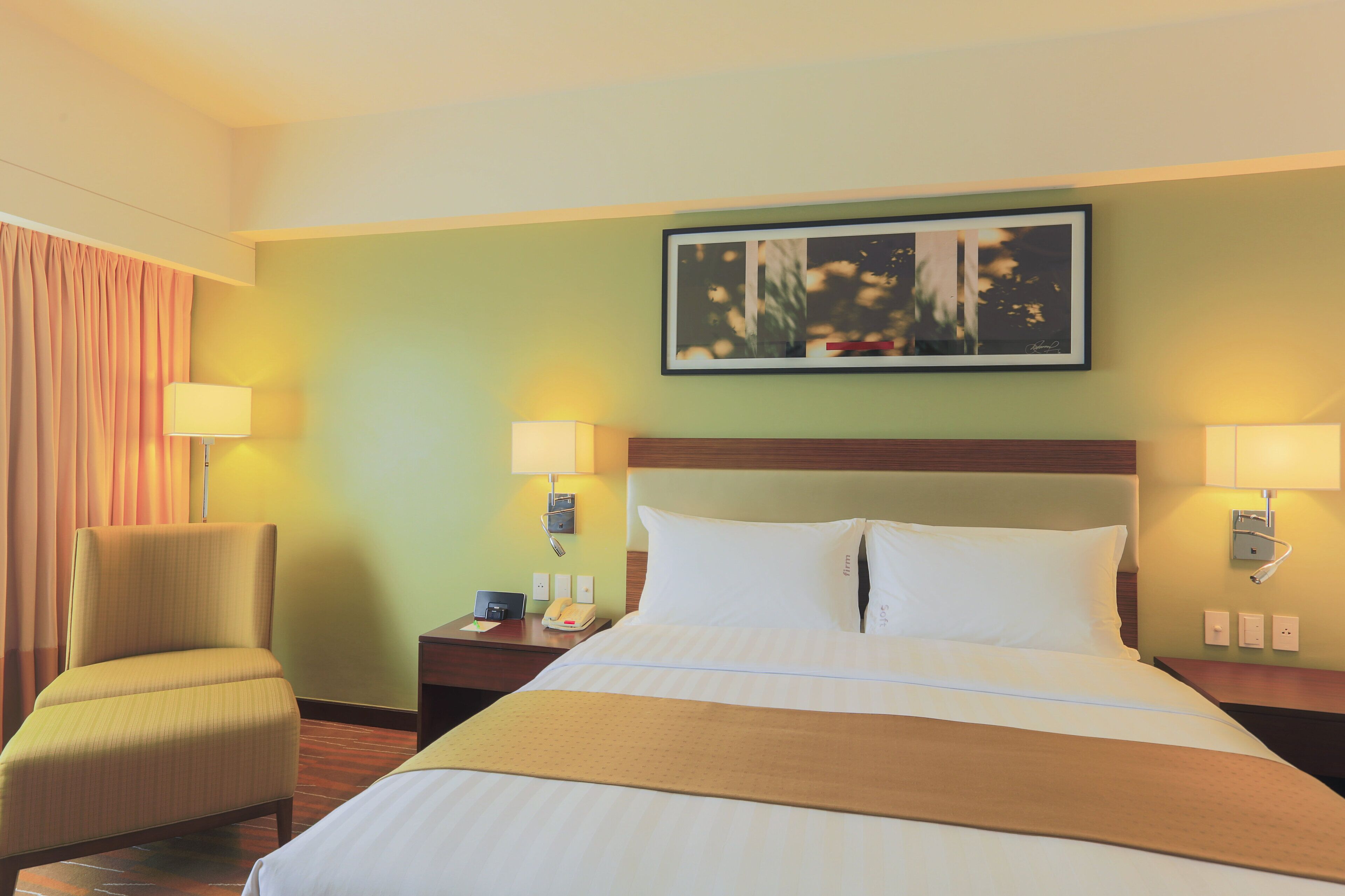 A hotel room with a bed, a chair, a nightstand, and a wall-mounted lamp.
