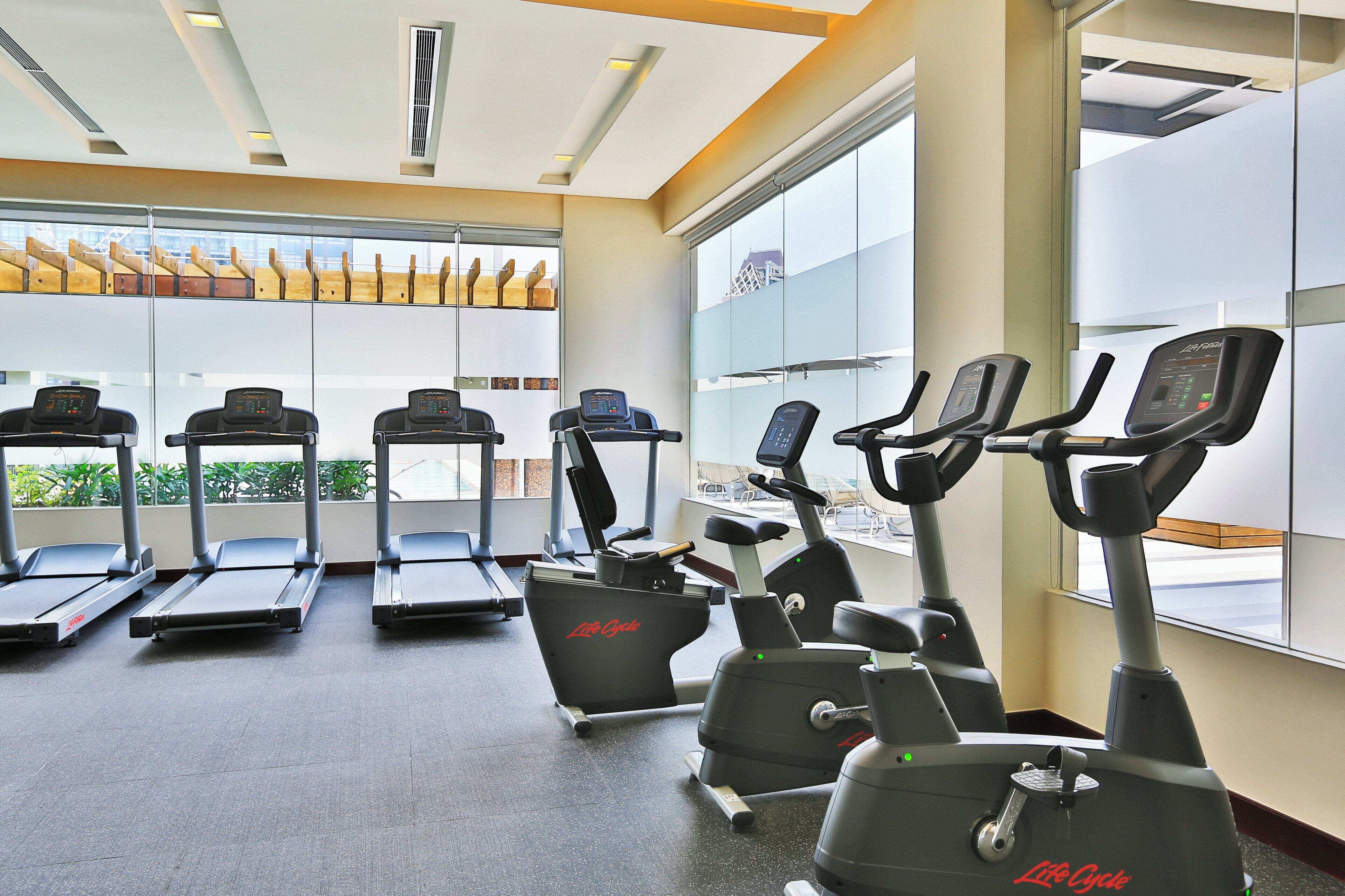 A gym with treadmills and elliptical machines, large windows, and a ceiling with recessed lighting.