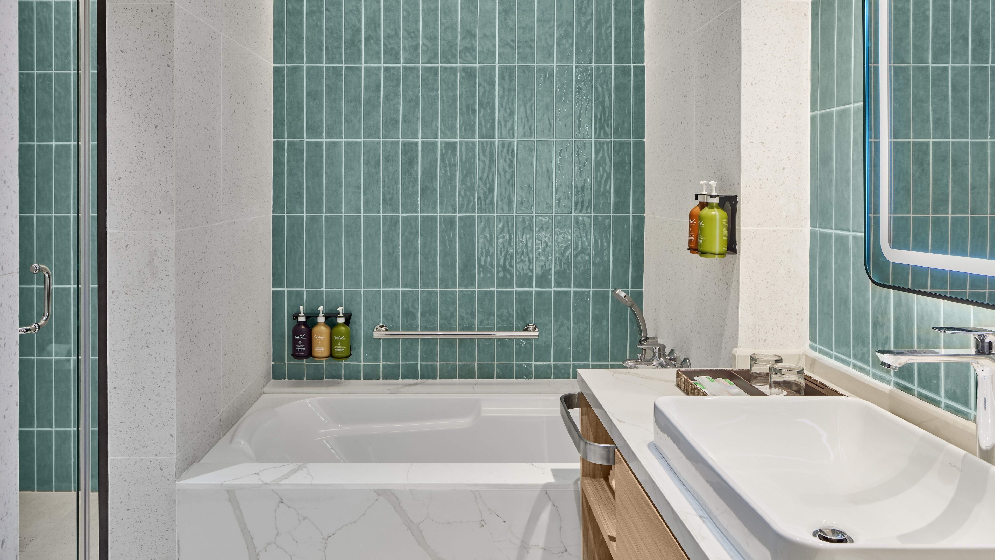Un baño con bañera, lavamanos y espejo. Hay tres botellas de champú en un estante.