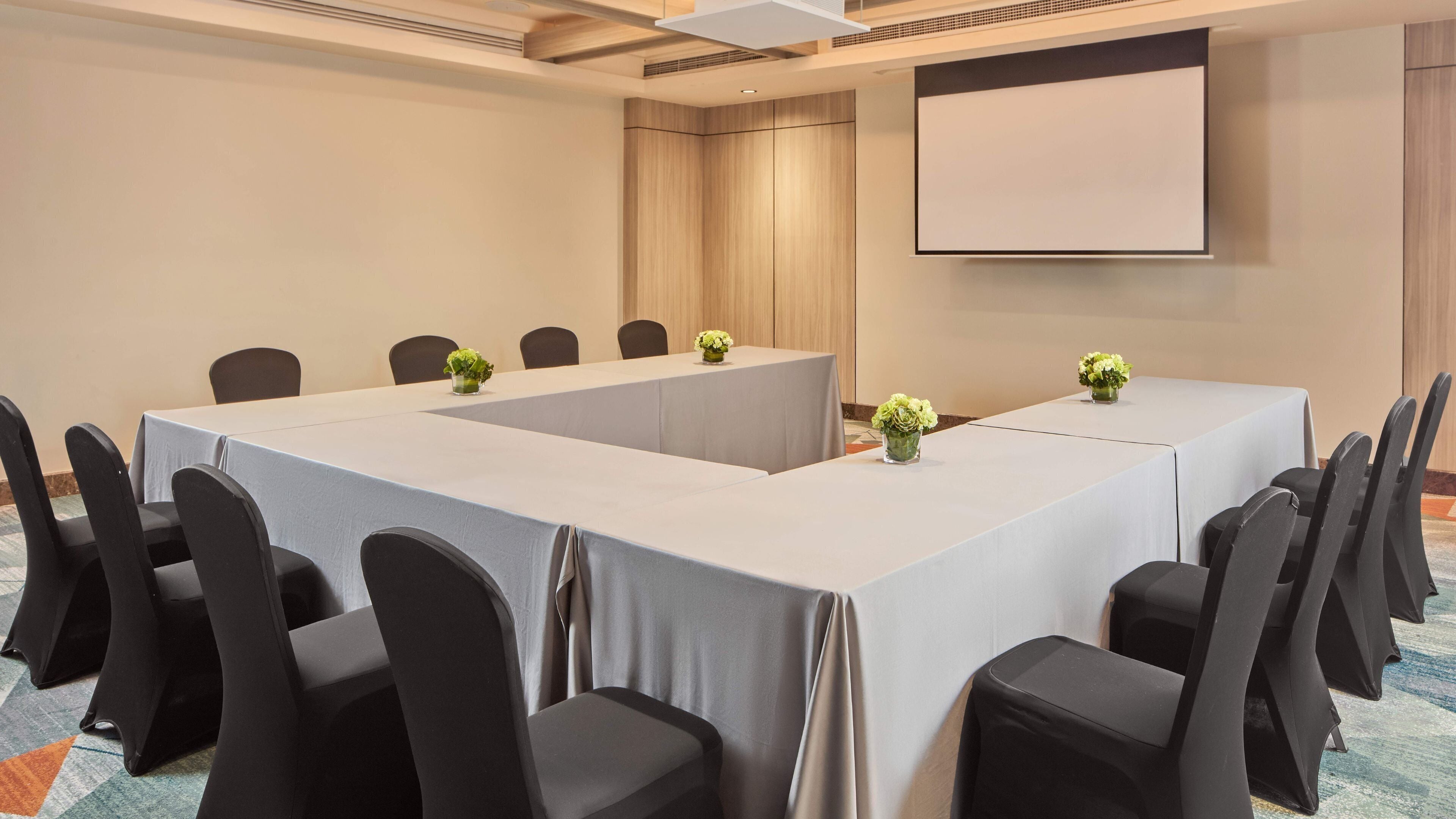Un salón de conferencias con una larga mesa, sillas negras y una pantalla de proyector.