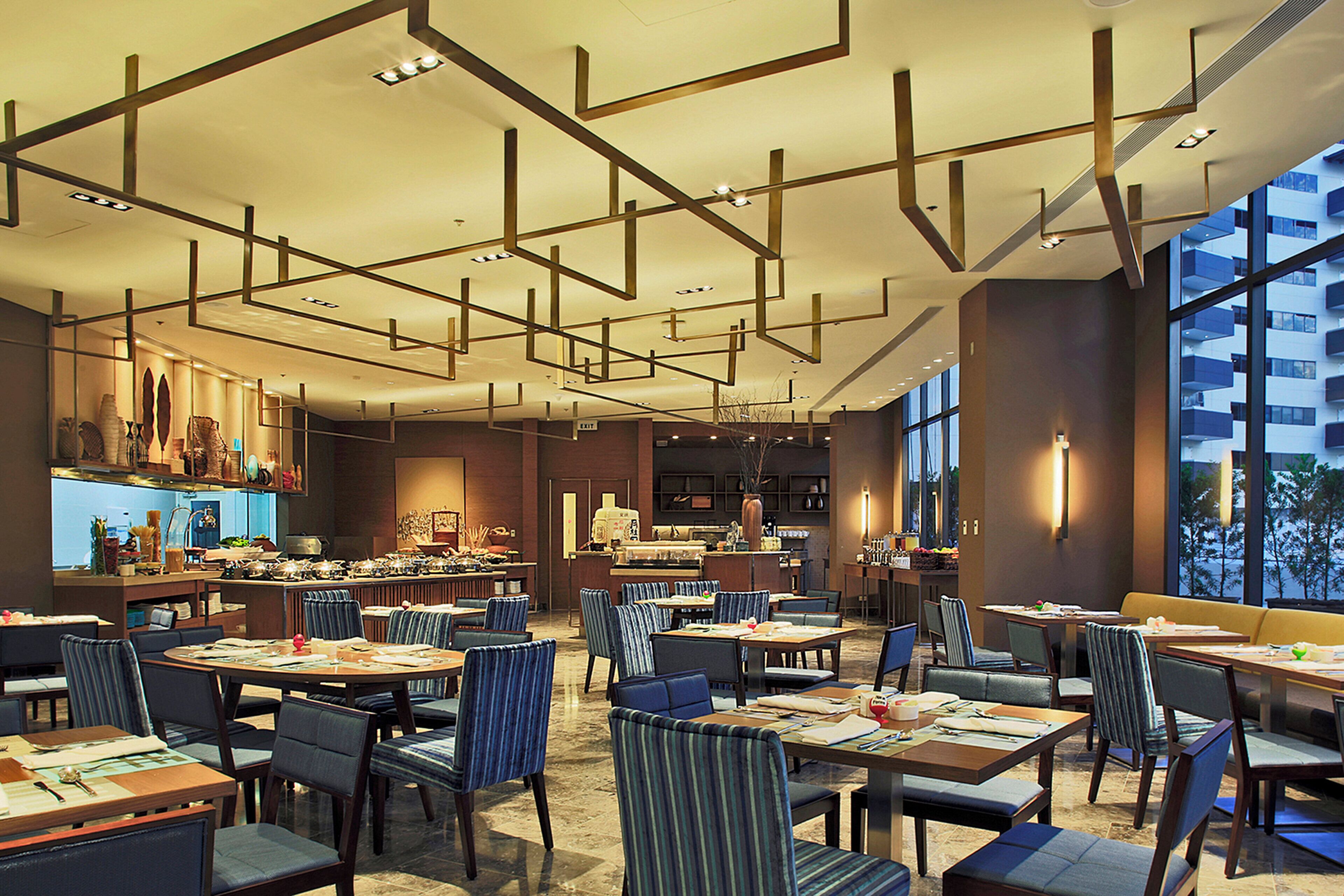 A modern restaurant interior with high ceilings, geometric ceiling design, and large windows offering views of the outside.
