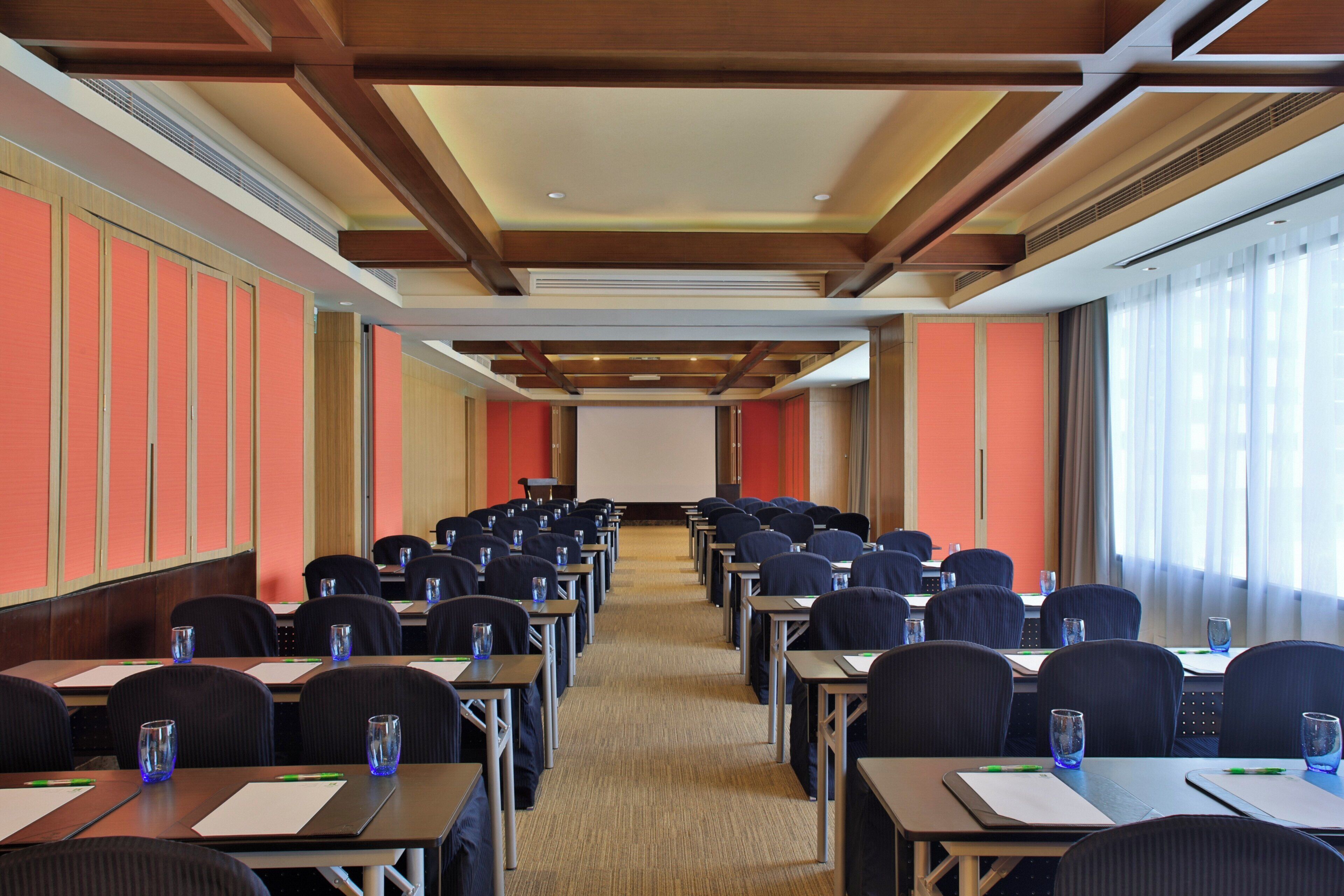 A conference room with rows of tables and chairs, a projector screen at the front, and large windows on one side.