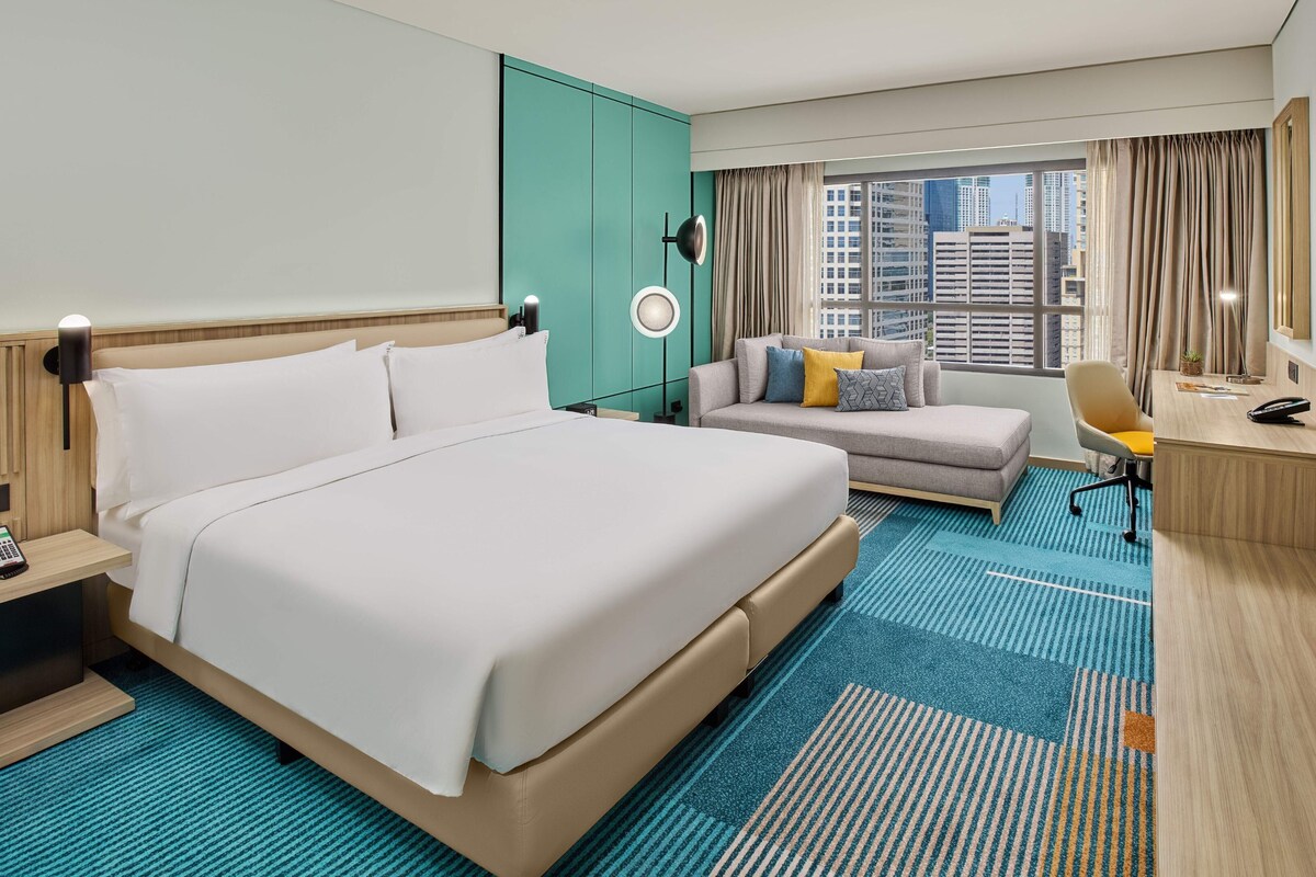 A hotel room with a large bed, a desk, a chair, a sofa, and a view of the cityscape.