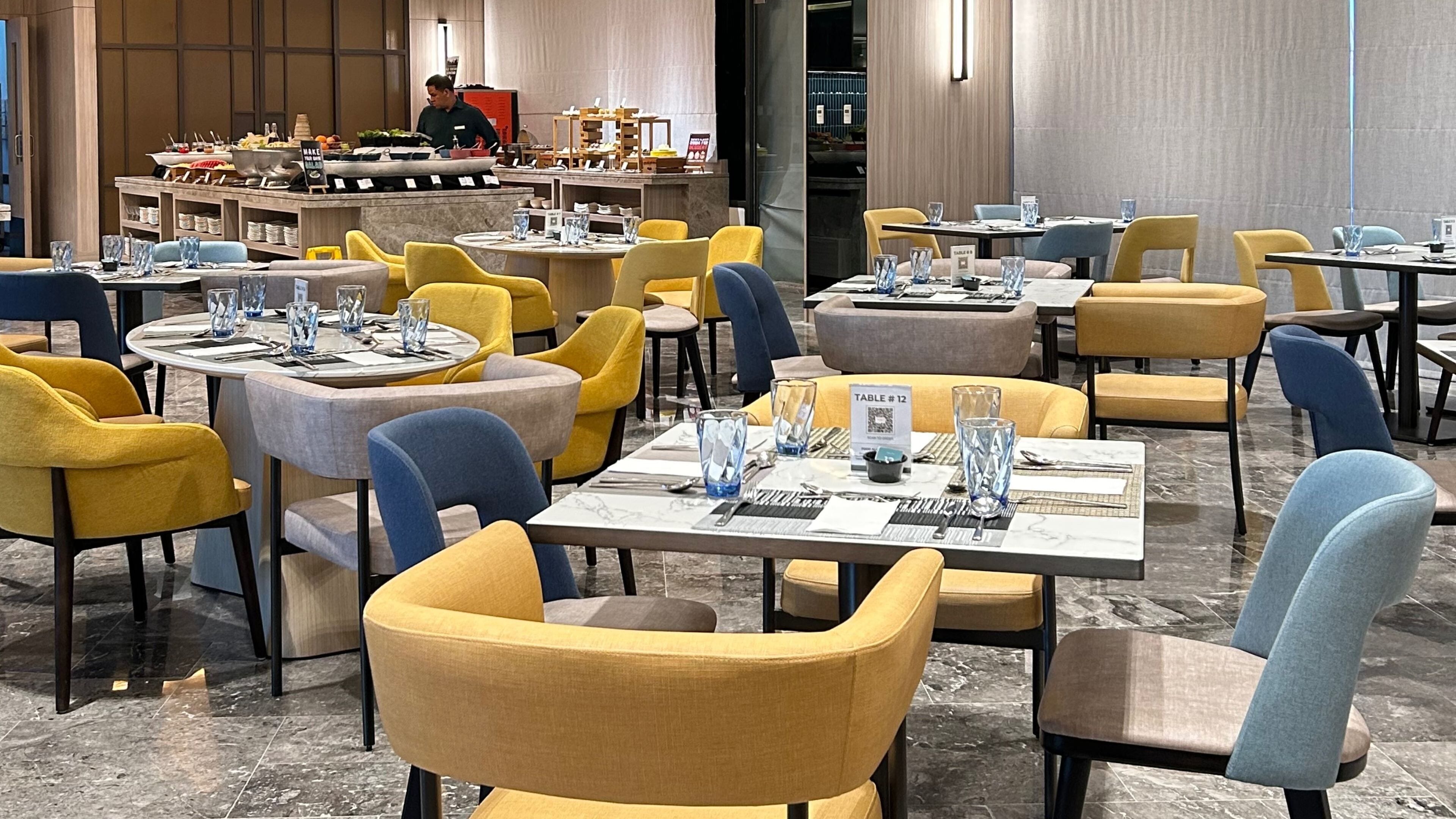 A restaurant with multiple tables and chairs, some with yellow upholstery. There are glassware and cutlery on the tables, and a person is visible in the background.