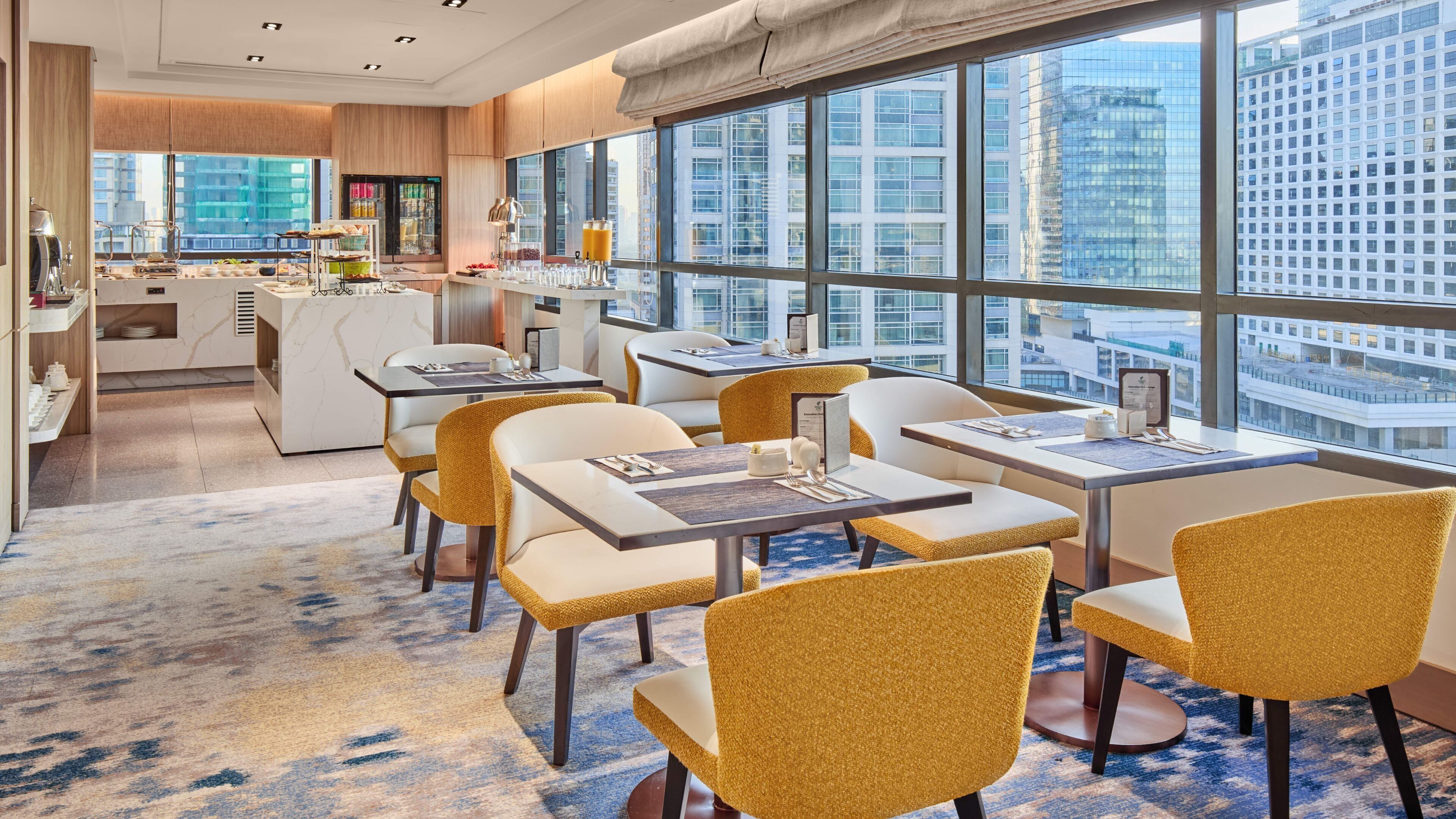 Comedor moderno de hotel con mesas y sillas, ventanas amplias con vistas a la ciudad y una cocina al fondo.