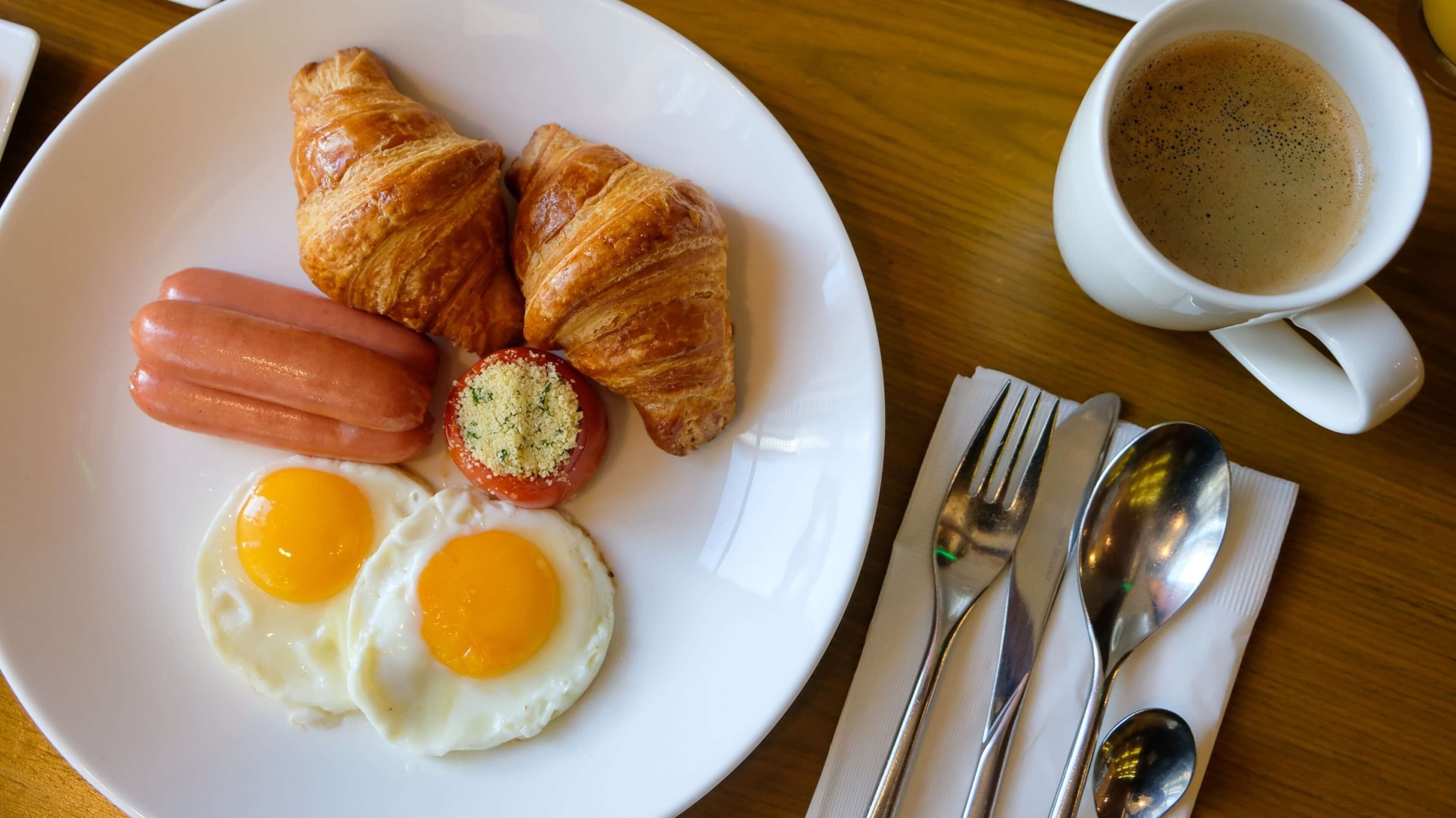 A plate with croissants, sausages, and eggs, a cup of coffee, and cutlery on a wooden table.