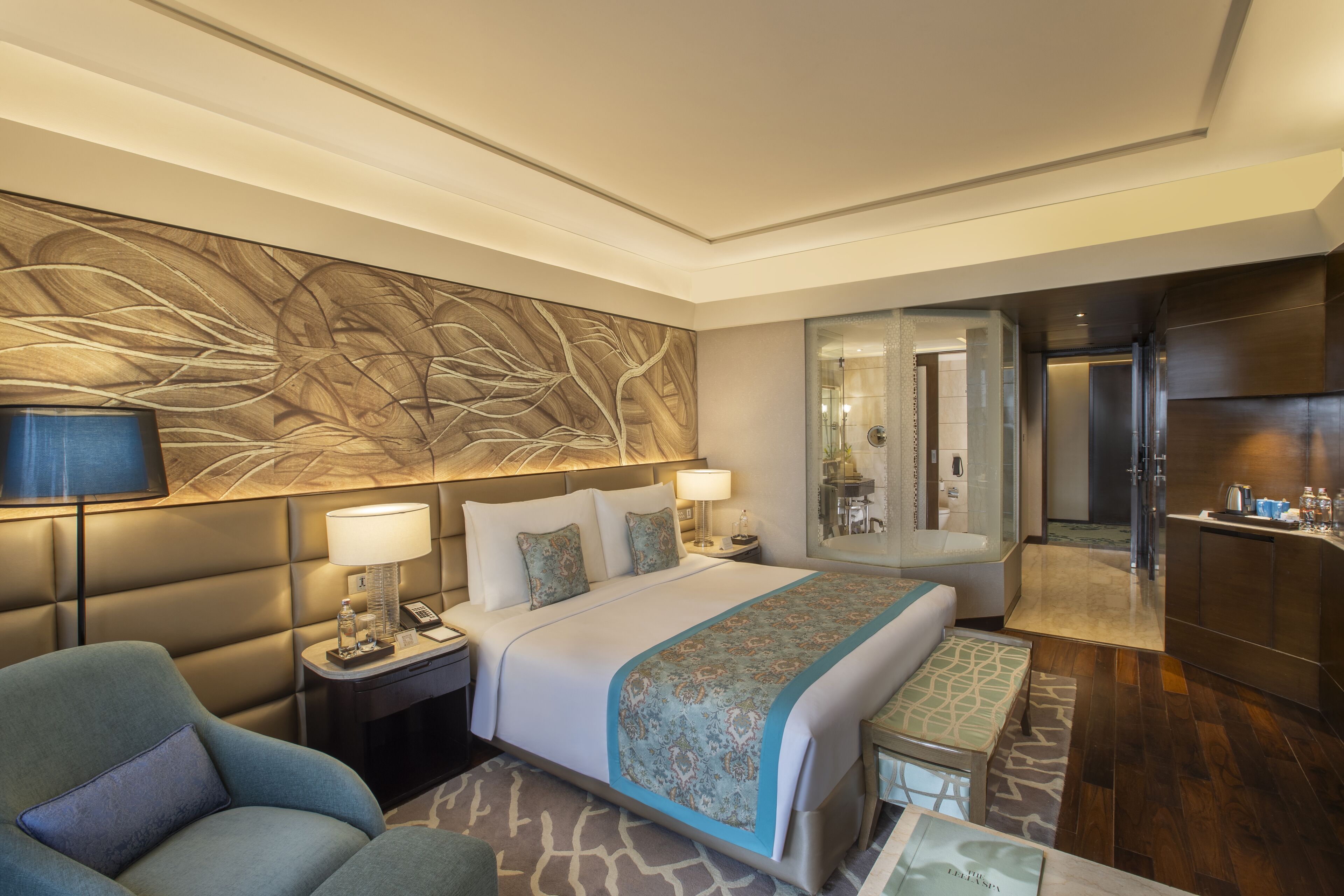 Habitación de hotel moderna con una cama grande, zona de estar con sofá, escritorio y baño visible a través de un vidrio.