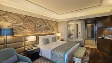 Une chambre d’hôtel moderne dotée d’un grand lit, d’un coin salon avec un canapé, d’un bureau et d’une salle de bain visible à travers une porte en verre.